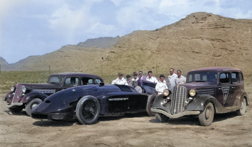 A group of men posing next to some classic cars.
