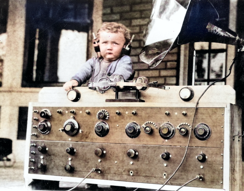 An image, taken in the mid-20th century, of a young boy playing with an old radio, while wearing big headphones.