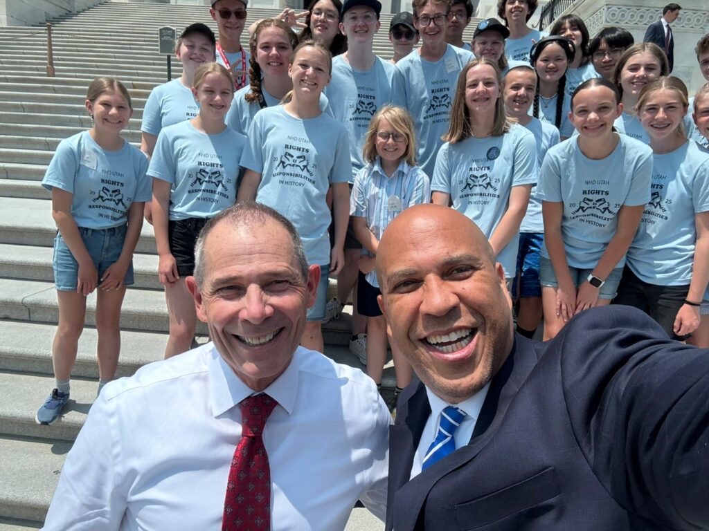 Sen. John Curtis (R, UT) and Sen. Cory Booker (D, NY) pose for a selfie with students from the NHD Utah program on the steps of the U.S. Capitol building.