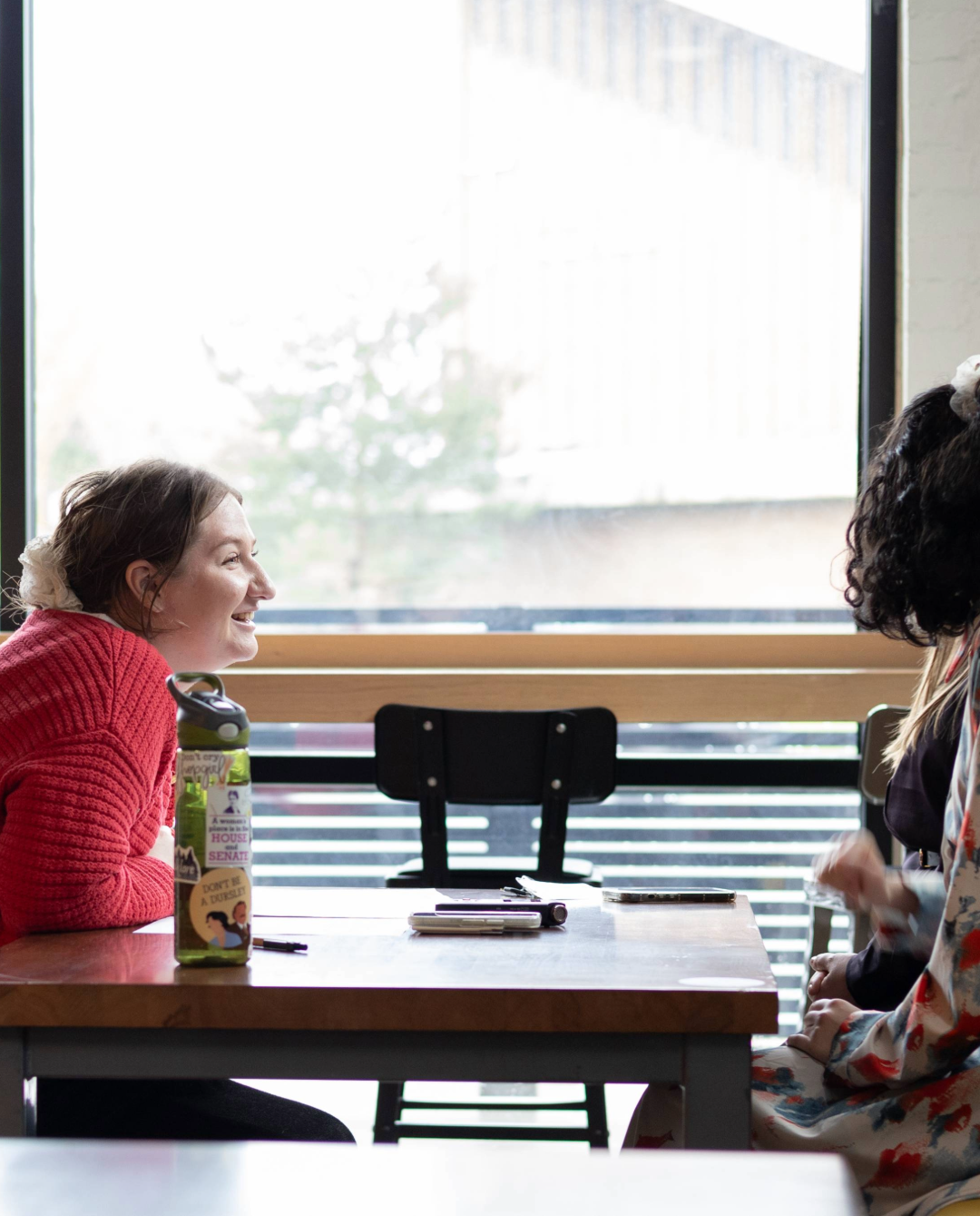 Two people sit across from each other at a table, talking by a large window.