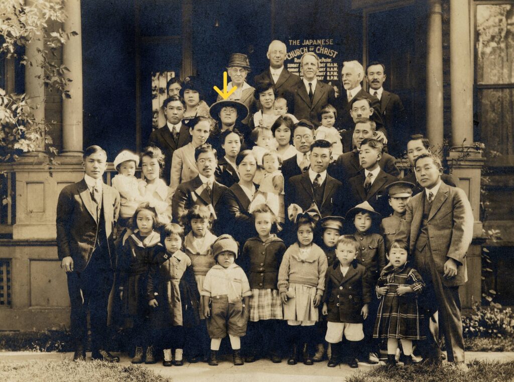 A large group of people poses in front of a building; one person is marked with a yellow arrow.