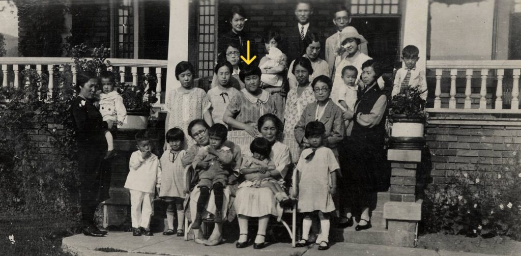 A group of people pose for a photo outside a house; an arrow points to one woman in the center.