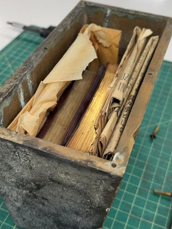 A weathered metal box contains old books, papers, and newspapers on a green cutting mat.